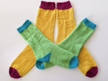 Vier bunte handgestrickte Socken in Gelb und Hellgrün mit Herz- und LOVE-Mustern auf hellem Hintergrund.