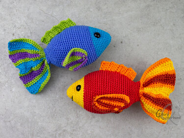 Zwei gehäkelte Amigurumi-Fische, einer blau-grün mit gestreiften Flossen, einer rot-gelb mit orange-roten Flossen auf grauem Untergrund