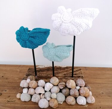 Three crochet shell ornaments in white, teal and light blue on black rods fixed into a wood block, displayed above a collection of small seashells.