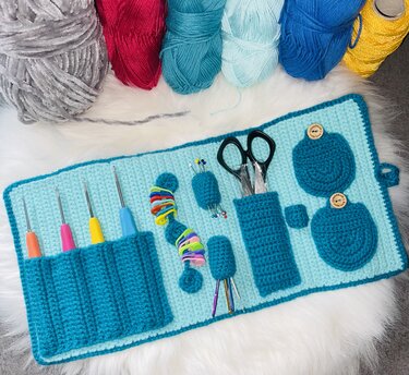 Gehäkelter Roll-Organizer in Türkis und Hellblau mit Häkelnadeln, Schere und Maschenmarkierern, umgeben von bunten Wollknäueln auf einem weißen, flauschigen Untergrund.