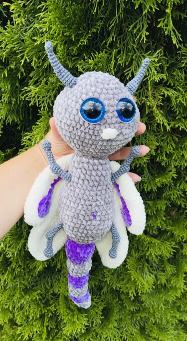 Gehäkelte Amigurumi-Motte in Grau, Weiß und Violett mit großen blauen Augen vor grünem Hintergrund