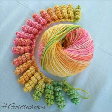 Mehrfarbiges Garnknäuel und gehäkelte Goldlöckchen-Locken in abgestuften Farben auf hellem Hintergrund.