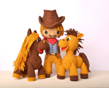 Amigurumi cowboy doll with brown hat and red bandana between two crocheted horses, one brown with a yellow mane and one golden with a brown mane.