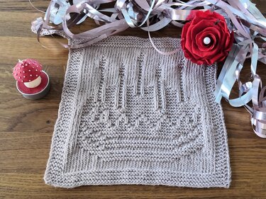 Quadratisches Strickstück in Creme‑Beige mit Relief-Kerzenmotiv, rosa Dekoband und roter Papierblume auf Holztisch.