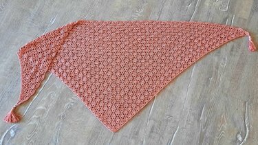 Dreieckiges lachsfarbenes Häkeltuch mit durchbrochenem Muschelmuster und Quasten auf Holzuntergrund.