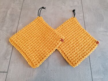 Zwei gehäkelte quadratische Topflappen in Senfgelb mit schwarzen Aufhängeschlaufen