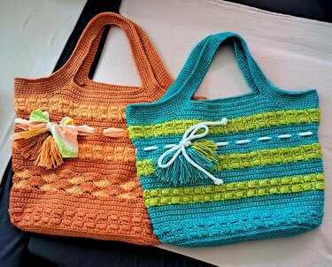 Zwei gehäkelte Shopper-Taschen, eine orange und eine türkisgrüne mit limettengrünen Streifen, jeweils mit Quaste und Tragegriffen.