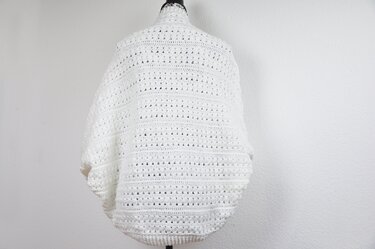 Back view of a white crocheted shrug on a dress form, showing openwork rows and ribbed hem