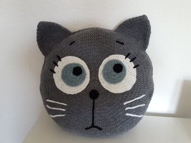 Gehäkeltes graues Katzenkopfkissen mit weißen Augenringen, hellblauer Iris, gestickter Nase und Schnurrhaaren.