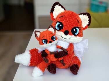 Two crocheted fox amigurumi, larger fox hugging smaller fox, orange and white with blue eyes and white ribbon, seated on a white surface.