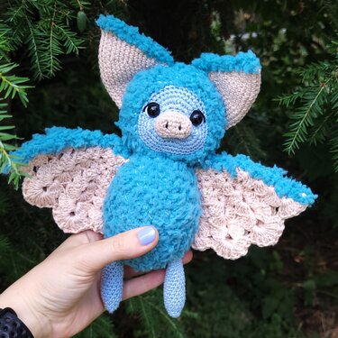 Hand hält türkisblaue gehäkelte Fledermaus-Amigurumi mit muschelförmigen, beigen Flügeln vor grünen Zweigen