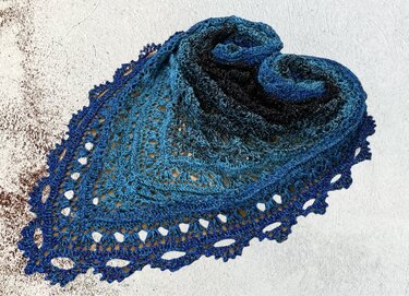 Folded blue ombre crocheted triangle shawl with openwork stitches and scalloped lace edge on a light textured surface.