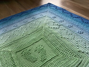 Gehäkelte Babydecke mit grün‑blauem Farbverlauf und Noppen- sowie Rippenstruktur auf Holzboden