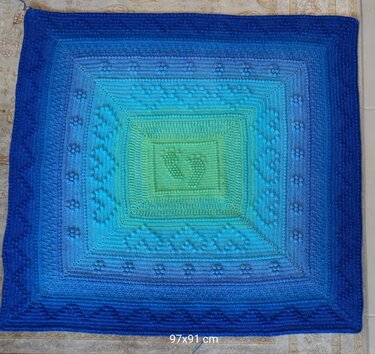 Quadratische gehäkelte Babydecke mit Ombré‑Farbverlauf und zentralen Babyfußabdrücken in Blau‑ und Grüntönen.