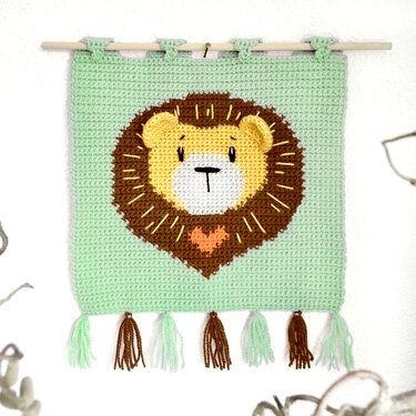 Handgehäkelter Wandbehang in Mintgrün mit gelbem Löwenkopf, brauner Mähne, orangefarbenem Herz und Quasten, an einem Holzstab aufgehängt.