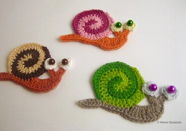 Drei gehäkelte Schneckenapplikationen mit bunten Spiralhäuschen und Perlenaugen auf hellem Untergrund