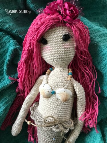 Gehäkelte Meerjungfrau-Amigurumi mit pinken Haaren, Muschel-Oberteil und hellgrauem Schwanz auf türkisfarbener Decke