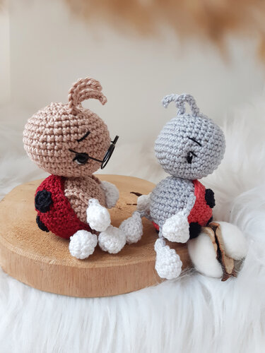 Two crocheted ladybug amigurumi with red spotted shells and white limbs; one wears small round glasses, displayed on a wooden slice.