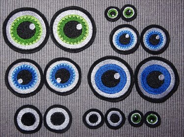 Mehrere gestickte, runde Augen-Applikationen in Grün und Blau sowie schwarz-weiße Varianten auf grauem, geripptem Stoff.