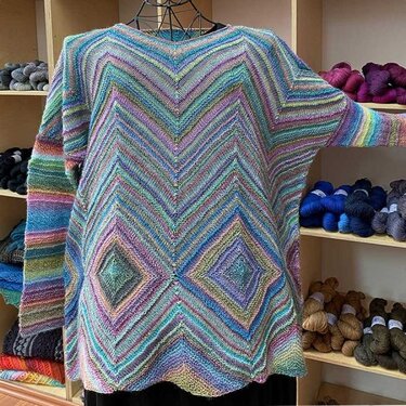 Front view of a multicolored knitted women's sweater with chevron stripes and central diamond motifs on a mannequin, surrounded by shelves of yarn.