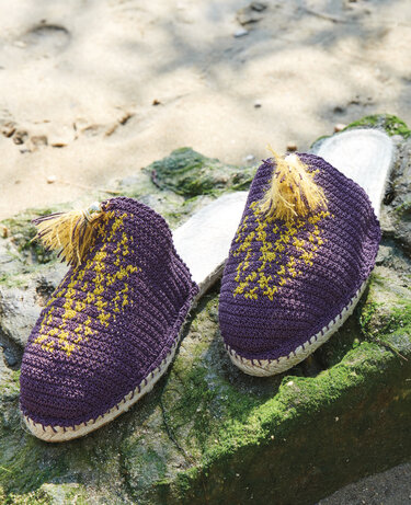 Paar gehäkelter violetter Pantoletten mit gelbem Muster und Quaste auf steinigem Untergrund