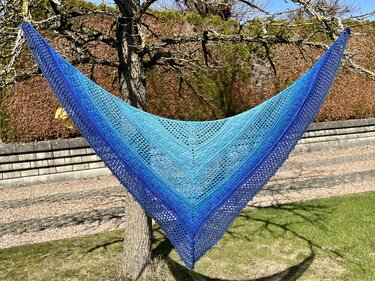 Gehäkeltes Dreieckstuch mit Blauverlauf und Spitzenmuster, aufgehängt an einem Baum