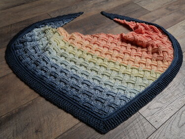 Gehäkeltes Dreieckstuch mit Ombre‑Farbverlauf (Blau, Gelb, Orange) im Korbgeflecht‑Muster und dunkelblauem Rand auf Holzboden
