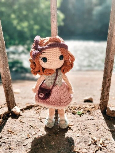 Gehäkelte Amigurumi‑Puppe mit kupferfarbenen Locken, rosa Rock, Stirnband und Umhängetasche, stehend im Freien