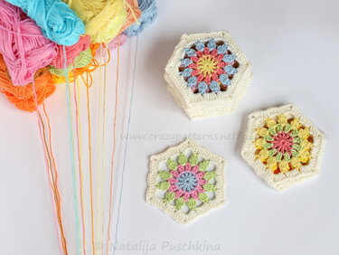 Drei gehäkelte Hexagon-Grannymotive in Pastelltönen neben Garnknäueln mit herabhängenden Fäden