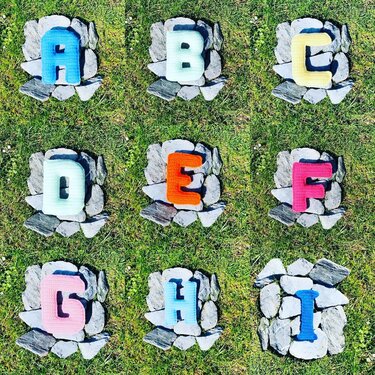 Overhead photo of nine crocheted block letters A to I in various colors, each centered on a circle of stones on grass