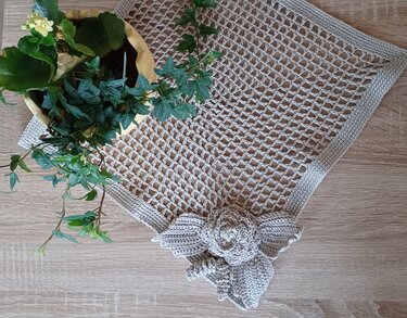 Hellbeiges, gehäkeltes Deckchen mit Gittermuster und gehäkelter Rose mit Blättern neben einer Topfpflanze auf hellem Holztisch.