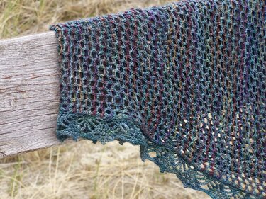 Nahaufnahme eines Strickstücks in Türkis-, Grün- und Violetttönen mit Lochmuster und dekorativem Rand auf einer Holzbank.