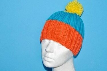 Orange-türkis gestreifte Strickmütze mit gelbem Bommel auf weißem Schaufensterkopf vor blauem Hintergrund.