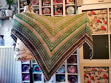 Crochet triangle shawl in green, beige, brown and dark gray gradient with textured stitch patterns, draped on a mannequin.