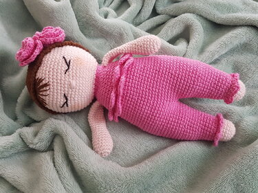 Gehäkelte Babypuppe in pinkem Outfit mit gestickten geschlossenen Augen und Blumenhaarband, liegend auf einer kuscheligen Decke