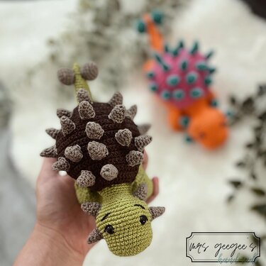 Hand holding olive green and brown crocheted Ankylosaurus amigurumi with spiked back; colorful dinosaur blurred in background.