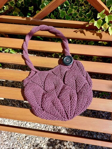Auberginefarbene, gehäkelte Henkeltasche mit erhabenem Strukturmuster, geflochtenem Henkel und rundem dunklem Knopf auf einer Holzbank.