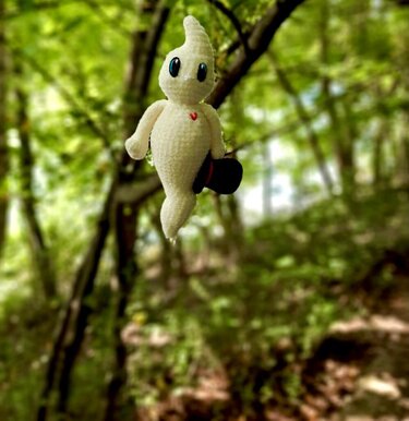 Weiß gehäkelter Amigurumi‑Geist mit blauen Augen, rotem Herz und schwarzem Zylinderhut vor grünem Wald