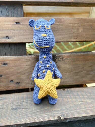 Handgehäkeltes blaues Amigurumi-Schlaftier mit geschlossenen Augen und gelben Sternen, hält einen großen gelben Stern vor Holz.