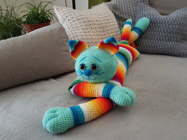 Gehäkeltes langes Katzenkissen mit bunten Regenbogenstreifen auf einem grauen Sofa