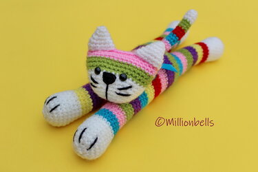 Striped amigurumi cat plush with rainbow colors, white face, black eyes and embroidered whiskers on a yellow background.