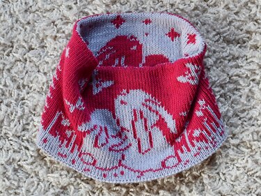 Red and light grey hand-knitted kids cowl with bunny and star Fair Isle motifs, placed on a cream shag rug.