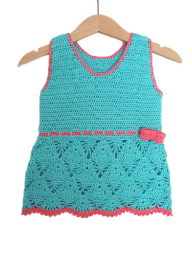 Gehäkeltes türkisfarbenes Kinder-Top mit Muschelmuster und rosa Kanten an einem Holzbügel