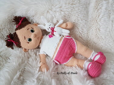 Gehäkelte Puppe mit Zöpfen, weißem Oberteil, rosa Höschen und kleinen rosa Schuhen, liegend auf hellem Fell, hält ein weißes Häschen