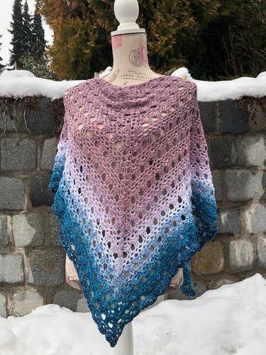 Gehäkeltes Dreieckstuch auf einer Schneiderpuppe mit Farbverlauf von Rosa über Weiß zu Blau, vor einer Steinmauer im Schnee.