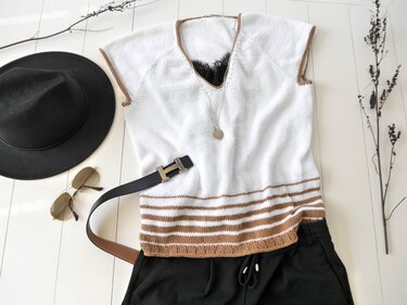 Weißes gestricktes ärmelloses Top mit braunen Streifen und V-Ausschnitt, flach gelegt neben einem Hut, einer Sonnenbrille und einem schwarzen G
rtel.