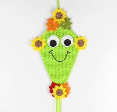 Lime green crocheted kite-shaped hanging decoration with big eyes, stitched smile and yellow-orange flower appliqués