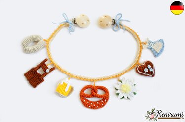 Handgehäkelte Schnullerkette mit bayerischen Anhängern (Brezel, Maßkrug, Edelweiß, Herz) an beige-orangefarbener Kordel und zwei Holzclips.