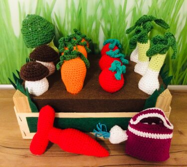 Mini-Gemüsegarten aus gehäkelten Karotten, Tomaten, Pilzen und Lauch in einer Holzkiste, daneben eine rote Chilischote, ein kleiner Pilz und ein lila Körbchen.