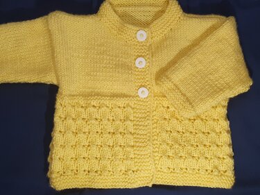 Gestrickte hellgelbe Babyjacke mit drei weißen Knöpfen und strukturiertem Saum auf dunkelblauem Untergrund